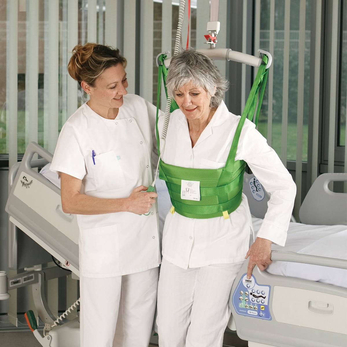 Klinikmitarbeiter, der einem Patienten mit einem Hillrom Deckenlifter und einer MasterVest beim Gehen in einem Krankenhauszimmer hilft